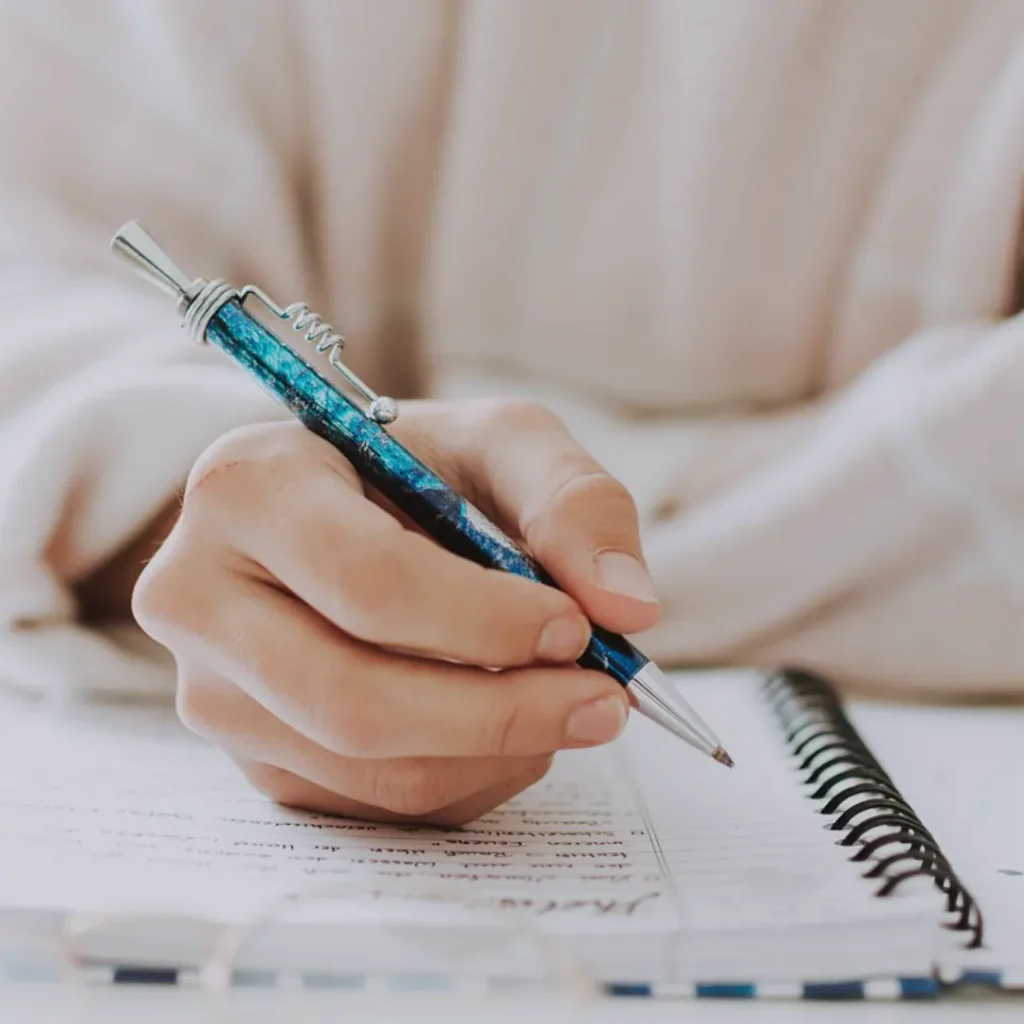 Das Bild zeigt eine rechte Hand, die einen Kugelschreiber über einem Blatt eines Notizbuches hält.
